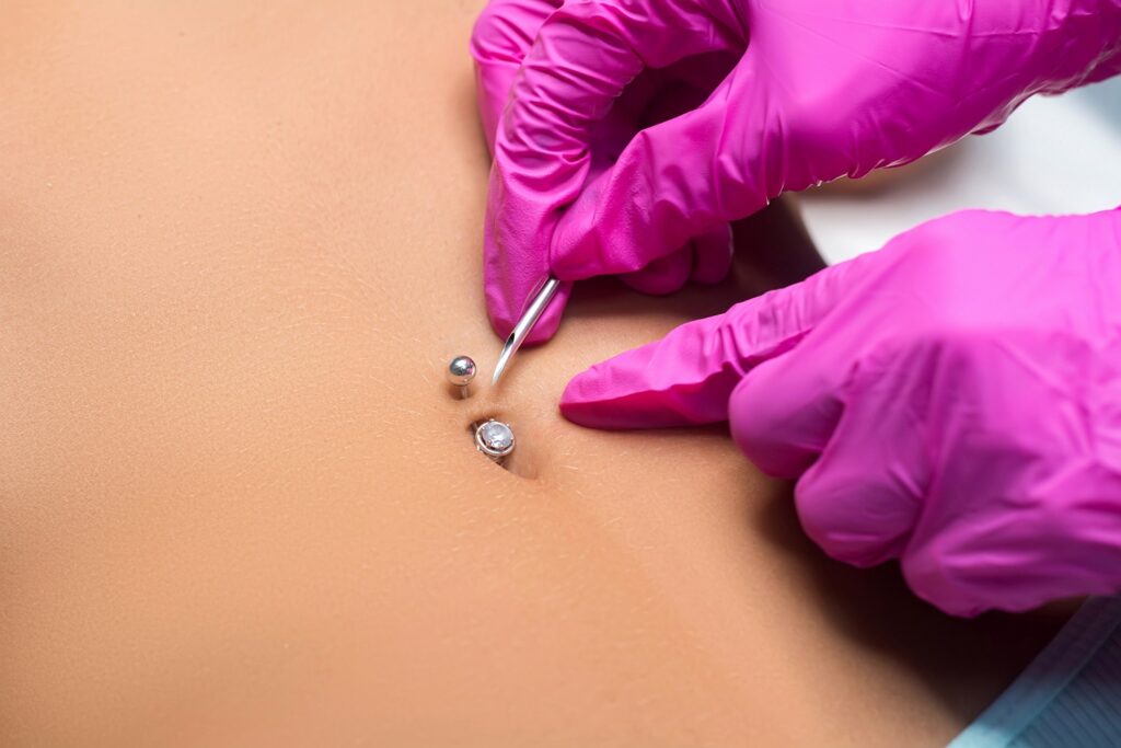 A belly button being pierced by a person with pink gloved hands.