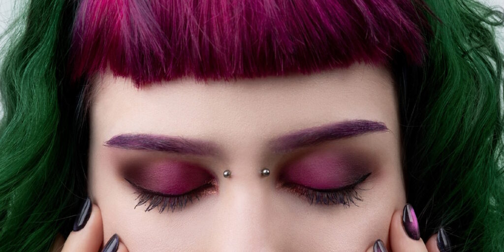 A woman with a lot of makeup and purple hair with a bridge piercing.