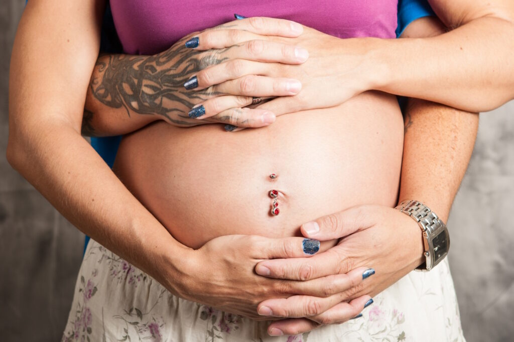A pregnant woman with a males hands around her belly that has a navel piercing.