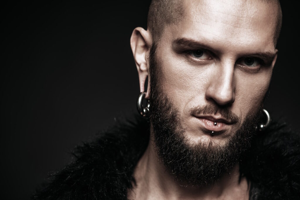 Portrait of a bearded man wearing a black fur coat with piercings in the ears and lips against a dark background.