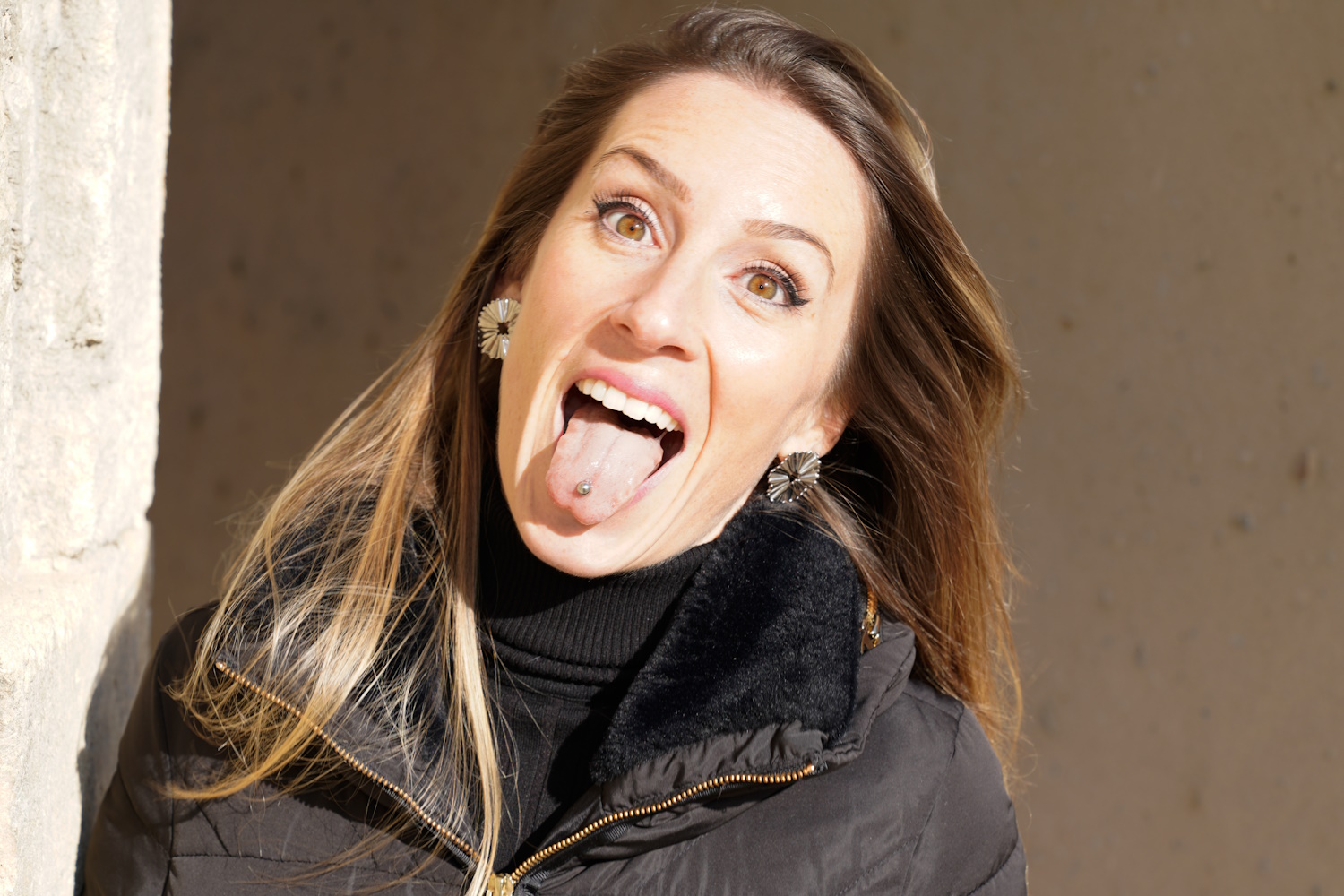Woman with a black shirt in a jacket with tongue piercing sticking her tongue out.